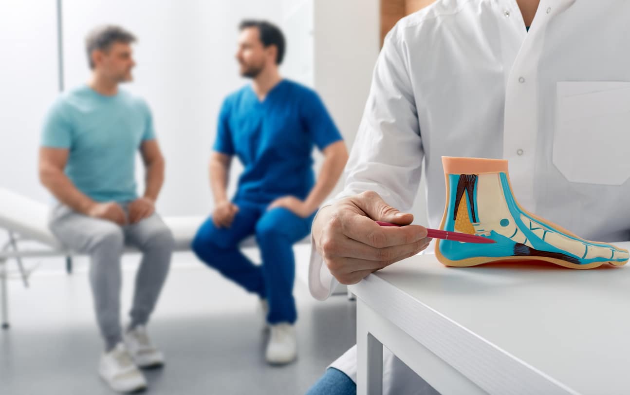A doctor pointing to a model of the inside of a foot with a patient and doctor in the background conversing on a patient’s table.
