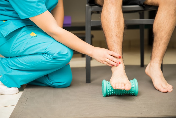 A female physical therapist assisting a male patient using a green foot roller to manage pain from plantar fasciitis.