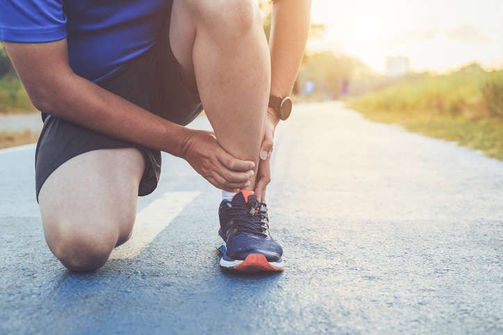 A man in a dark blue shirt, black shorts, and black sneakers sits holding his ankle in pain from tarsal tunnel syndrome.