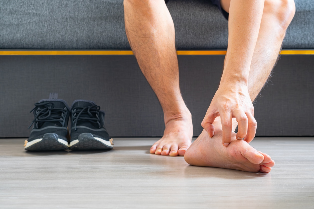 A man is examining his bare feet for signs of fungal infection with his black sneakers next to them.